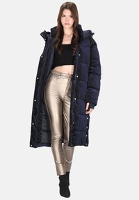 Długi granatowy puchowy płaszcz z kapturem, z przeszywanym wzorem i złotymi elementami; zestawiony z dopasowanymi metalicznymi złotymi spodniami.