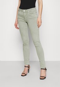 Jeans ajustados verde claro con una textura suave, que cuentan con bolsillos frontales y cremallera. Combinados con sandalias negras de tacón alto con tiras.