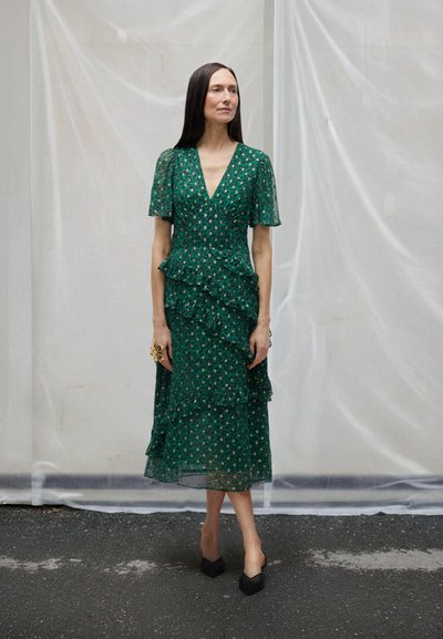 Žena s dlouhými tmavými vlasy, oblečená v zelených květovaných midi šatech s volánky a černými špičatými podpatky, stojící před bílým pozadím.