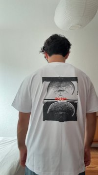 Camiseta blanca de manga corta con un diseño gráfico negro de círculos concéntricos en la parte trasera, con texto rojo debajo del gráfico.