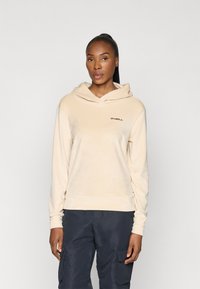 Beżowy polarowy hoodie z kieszenią kangurzą z przodu, regulowanym kapturem ze sznurkiem i haftowanym logo na klatce piersiowej. Noszony z ciemnymi spodniami.