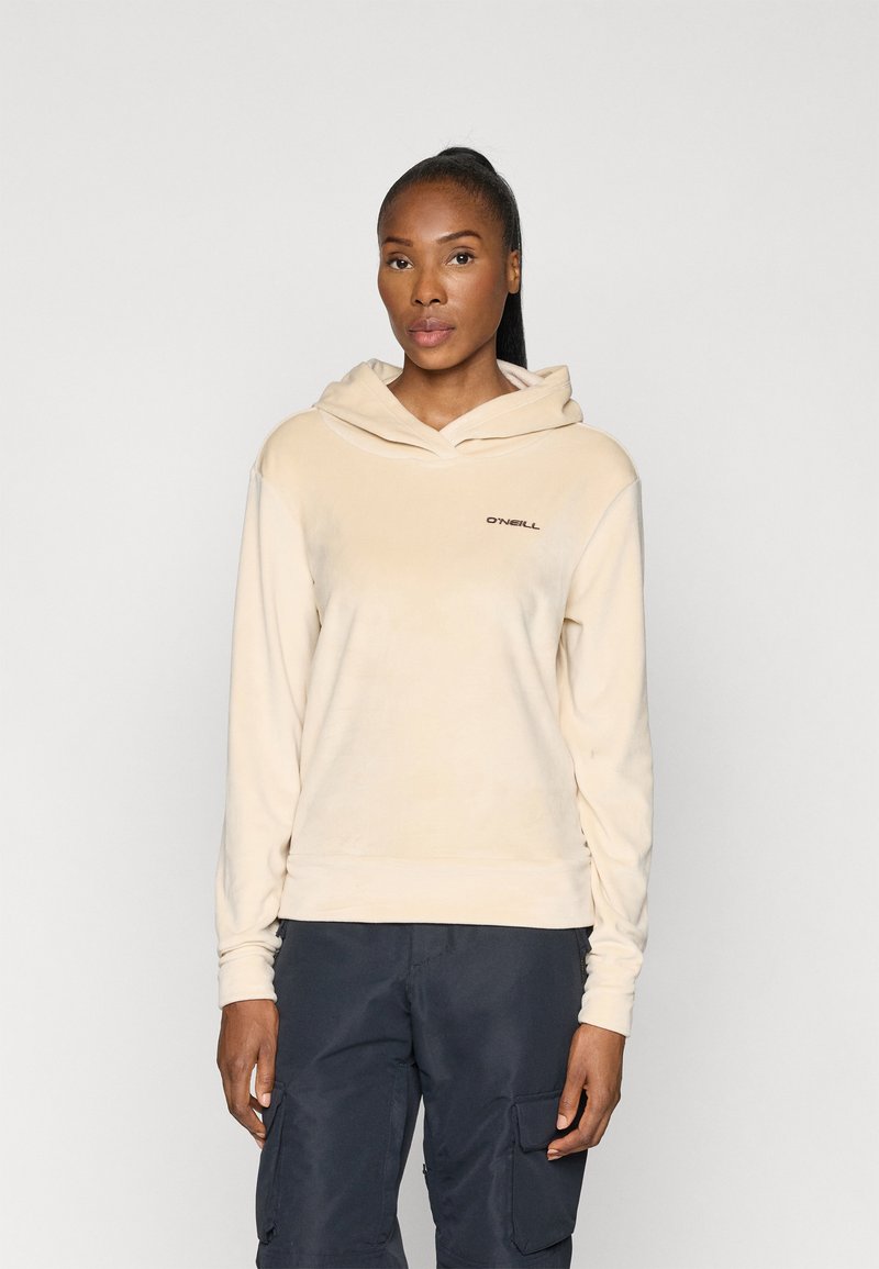 Beżowy polarowy hoodie z kieszenią kangurzą z przodu, regulowanym kapturem ze sznurkiem i haftowanym logo na klatce piersiowej. Noszony z ciemnymi spodniami.
