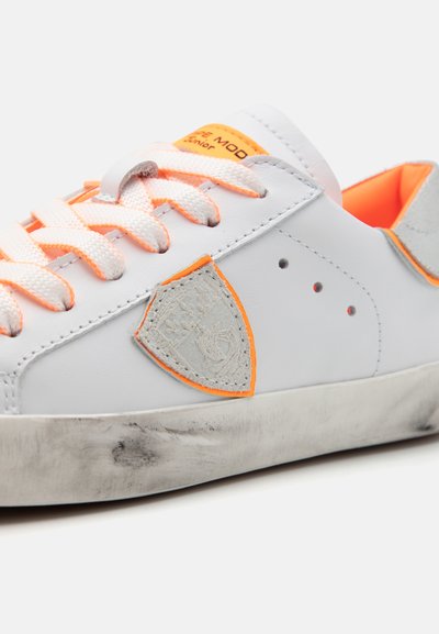 Białe skórzane sneakersy z pomarańczowymi akcentami i szarym emblematem. Posiadają lekko kontrastującą teksturowaną podeszwę, różowe sznurówki oraz minimalne perforacje.