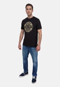 Czarny bawełniany T-shirt z okrągłym nadrukiem z napisem "WORK PETROL DRIVEN WEAR" w beżowym kolorze, w połączeniu z jasnoniebieskimi dżinsami i granatowymi sneakersami.