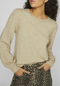 Bežs adīts džemperis ar garām piedurknēm un apaļu kaklu; uzvilkts virs melna un bežīga leopardu raksta svārkiem, kuros ir sānu kabatas.