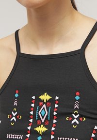 Musta toppi, jossa on värikkäitä brodeerattuja kuvioita punaisesta, keltaisesta, sinisestä ja valkoisesta. Säädettävät olkahihnat ja muotoiltu malli.