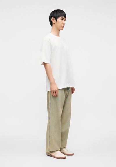 Λευκό κοντομάνικο μπλουζάκι με ένα μικρό λογότυπο, συνδυασμένο με ανοιχτό πράσινο παντελόνι φαρδύ με ρίγες και μπεζ αθλητικά παπούτσια με καφέ σόλες.