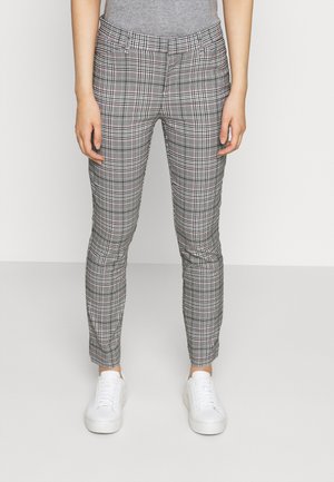 Žena v úzkých kostkovaných kalhotách s růžovými akcenty, šedém topu a bílých teniskách, stojící před jednobarevným světlým pozadím.