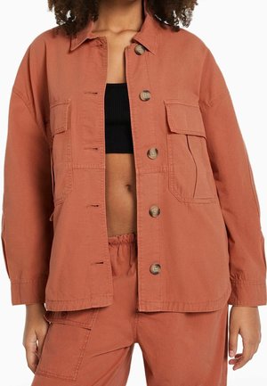 Femme portant une veste à boutons de couleur rouille et un pantalon assorti avec un haut court noir laissant voir le ventre, sur fond blanc.