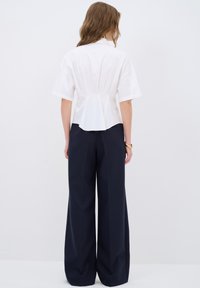 Camisa blanca de manga corta con detalles fruncidos en la parte posterior, combinada con pantalones de talle alto y pierna ancha en color marino. Diseño minimalista.