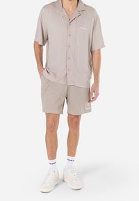 Světlé béžové tričko s krátkými rukávy, výstřihem s výřezem a zapínáním na knoflíky vpředu, doplněné světlými béžovými síťovanými šortkami s jemným vzorem a logem.