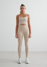 Top corto atletico beige con spalline sottili abbinato a leggings a vita alta, entrambi in tessuto liscio. Scarpe da ginnastica bianche con accenti grigi.