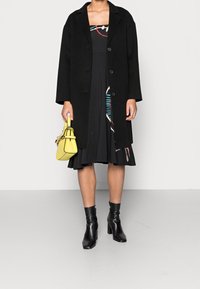 Musta villakangastakki, jossa on suuret napit, kerroksittain mustan mekon päällä, jossa on värikkäitä geometrisia kuvioita. Keltainen käsilaukku ja mustat korkokengät.