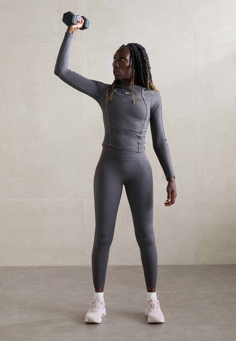 Haut de sport gris avec col montant, leggings ajustés assortis et baskets rose clair. La personne tient un haltère au-dessus de la tête. Texture lisse.