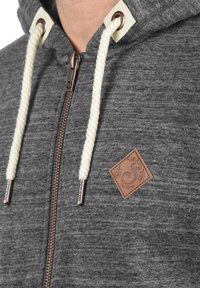 Grijze rits hoodie gemaakt van zachte stof, met een bronzen rits, crèmekleurige trekkoorden en een bruin leren logo label op de borst.