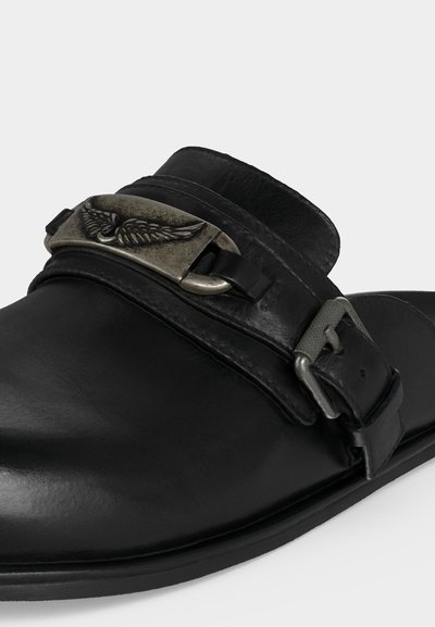 Μαύρο δερμάτινο παπούτσι με μεταλλικό έμβλημα με φτερά στο λουράκι και ασημένια αγκράφα στο πλάι, εμφανίζεται σε λευκό φόντο.