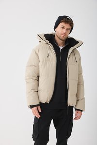 Bărbat îmbrăcat cu o geacă puffer bej, hanorac negru, pantaloni cargo negri și o căciulă tricotată neagră, stând în fața unui fundal alb simplu.