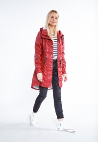 Impermeabile rosso con zip frontale, cappuccio regolabile e vita con cordino. Abbinato a una maglietta a righe, jeans neri e sneakers bianche.