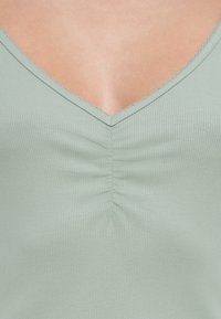 Πράσινο- μέντας ριπ τοπ με βαθιά λαιμόκοψη σε σχήμα V και πιέτες στο κέντρο. Ορατό υφή ύφασμα και ραμμένα άκρα.