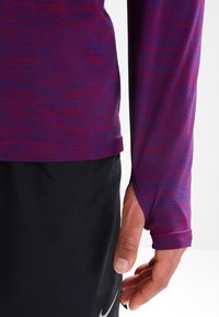 Długi rękaw sportowa bluzka w purpurowo-czerwonym melanżowym wzorze, wykonana z lekkiego materiału. Posiada dopasowany krój z otworami na kciuki.