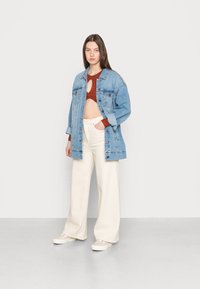 Casaco de ganga em azul claro, de corte oversized. Top curto castanho com recorte circular e calças largas em creme. Sapatilhas bejes completam o look.