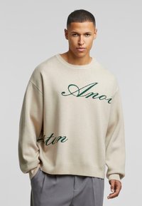 Beige oversized trui van gebreid materiaal met groene cursieve tekst "Amor" en "Texton" op de voorkant, met een ronde halslijn en geribbelde mouwen.