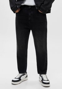 Personne portant un jean noir, des baskets blanches et noires, les mains dans les poches, avec une partie d'une veste noire visible.