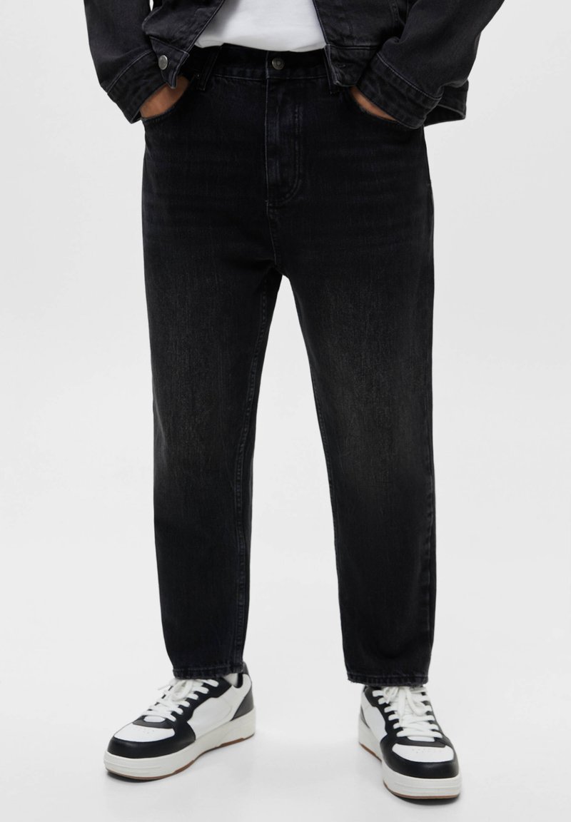 Personne portant un jean noir, des baskets blanches et noires, les mains dans les poches, avec une partie d'une veste noire visible.