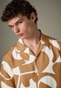 Trumpa rankovė, ruda ir balta abstraktaus rašto, lengva audinys, priekiniai mygtukai, išskleistas apykaklis, lygi tekstūra.