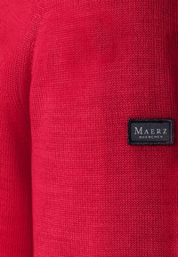 Suéter de punto rojo con una tela texturizada. Una etiqueta de logo negra con texto blanco es visible en la manga.