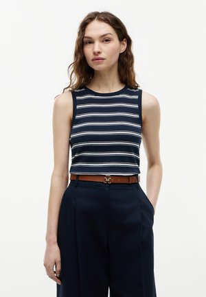 Γυναίκα με κυματιστά καστανά μαλλιά, που φοράει μπλε σκούρο και λευκό ριγέ αμάνικο τοπ, μπουσουλισμένο σε ψηλόμεσα μπλε σκούρα παντελόνια με καφέ ζώνη.