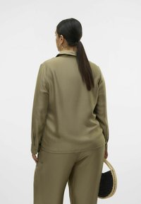 Olivgrön, strukturerad blus med krage och långa ärmar, matchad med byxor i samma färg. Modellen håller en liten, flingad svart handväska.