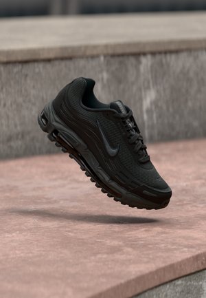 Czarny sportowy but Nike Air Max zawieszony nad kamienną powierzchnią z rozmytym kamiennym murem w tle.