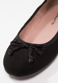 Ballerine plate en daim noir avec un nœud décoratif en corde torsadée sur l'avant et une semelle intérieure rose pâle, photographiée sur fond blanc.