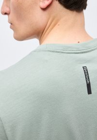 Άνδρας που φοράει ανοιχτοπράσινο t-shirt Reebok, θέα του λαιμού και του άνω μέρους της πλάτης με ορατό λογότυπο της μάρκας κοντά στον γιακά.