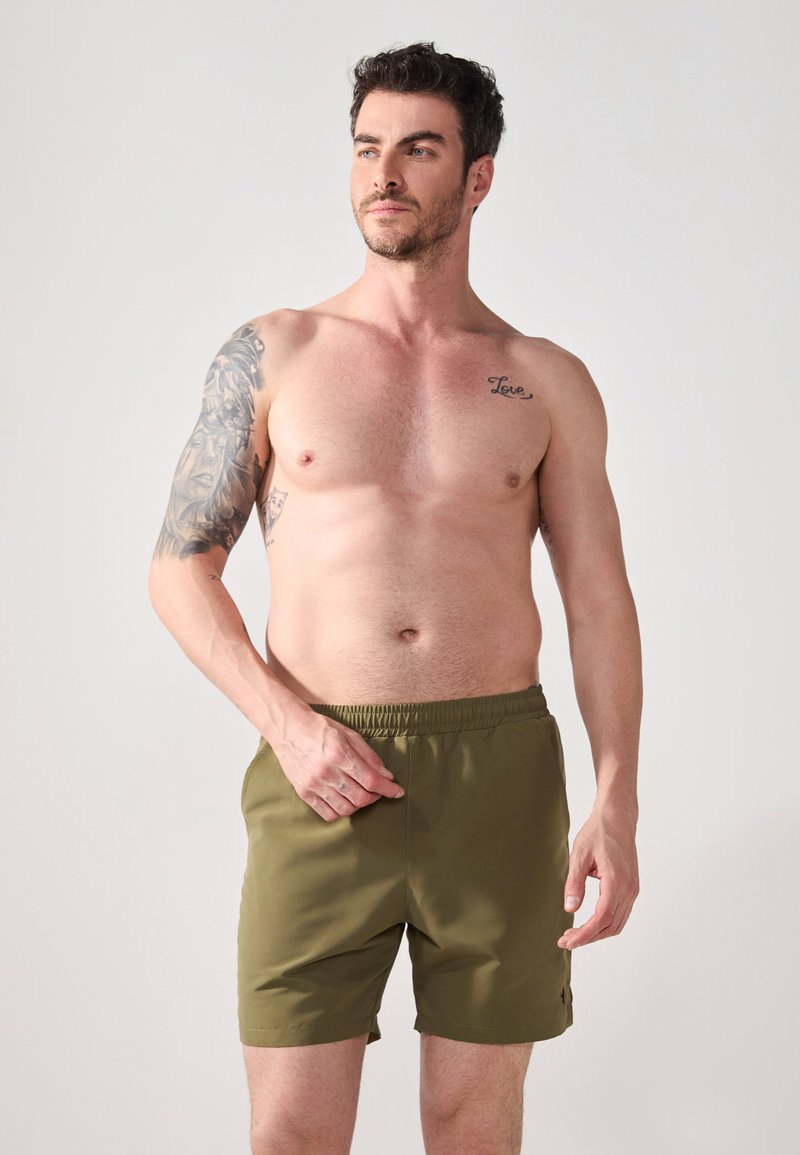 Khaki swim shorts with an elastic waistband and side pockets, made from a smooth, lightweight fabric, modeled by a shirtless man.