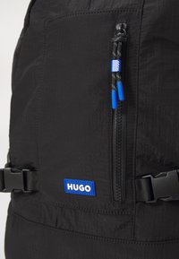 Sac à dos noir avec une surface texturée, fermeture éclair centrale avec des accents bleus, et un grand patch logo "HUGO" bleu. Comprend des bretelles réglables.