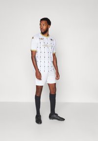 Vitt fotbollskit med svart geometrisk mönstring, guldaccent och "Versailles"-logotyp. I kombination med svarta shorts och matchande strumpor.