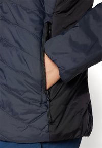 Marinblå vadderad jacka med en zippered ficka, framträdande kviltat mönster, slät textur och kontrasterande svart tyg på den nedre halvan.