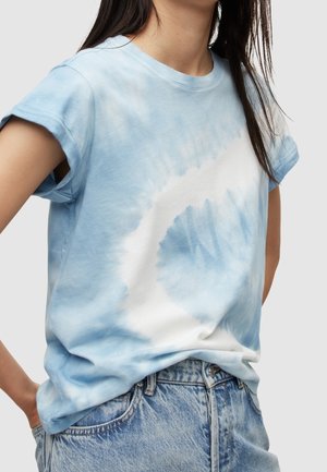 Mujer con camiseta tie-dye azul y blanca y vaqueros vaqueros azul claro, de pie con las manos en los bolsillos contra un fondo liso.