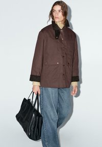 Giacca marrone con colletto in velluto a coste, due tasche frontali, indossata sopra un maglione crema, abbinata a jeans blu larghi e una borsa a tracolla nera.