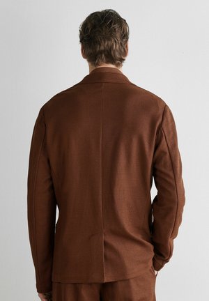 Mann mit kurzem welligem Haar, trägt eine braune Anzugjacke und passende Hose, steht vor einer schlichten weißen Wand.