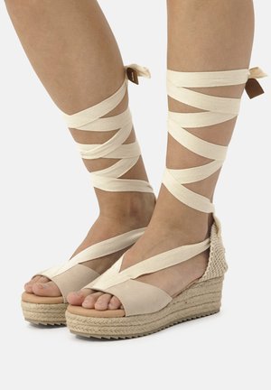 Sandali beige con zeppa e fascette in tela avvolte attorno alle caviglie, caratterizzati da una suola in juta texturizzata e un design open-toe. Accento marrone vicino alla caviglia.