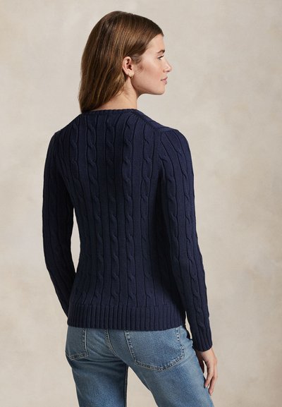 Kvinde med brunt hår iført en marineblå kabelstrikket sweater og lyseblå jeans, stående lidt drejet til siden mod en neutral baggrund.