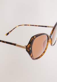 Sköldpaddsmönstrade solglasögon med bruna linser, bredare rundad båge, guldaccents på skalmarna och texturerad yta.