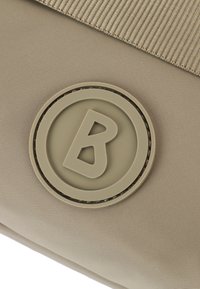 Borsa in tessuto beige con un logo circolare in rilievo con una "B" stilizzata al centro. Superficie texturizzata, con una tracolla piatta sopra.