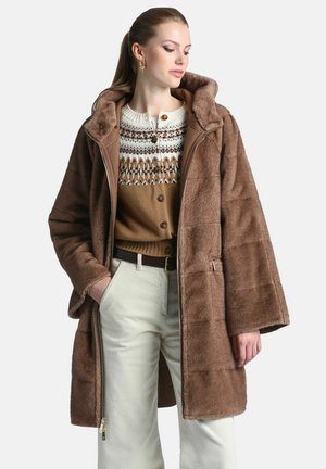 Femme portant un manteau à capuche en fausse fourrure marron, un cardigan marron à motifs, un pantalon blanc et des boucles d’oreilles dorées, regardant sur le côté avec une main dans la poche.