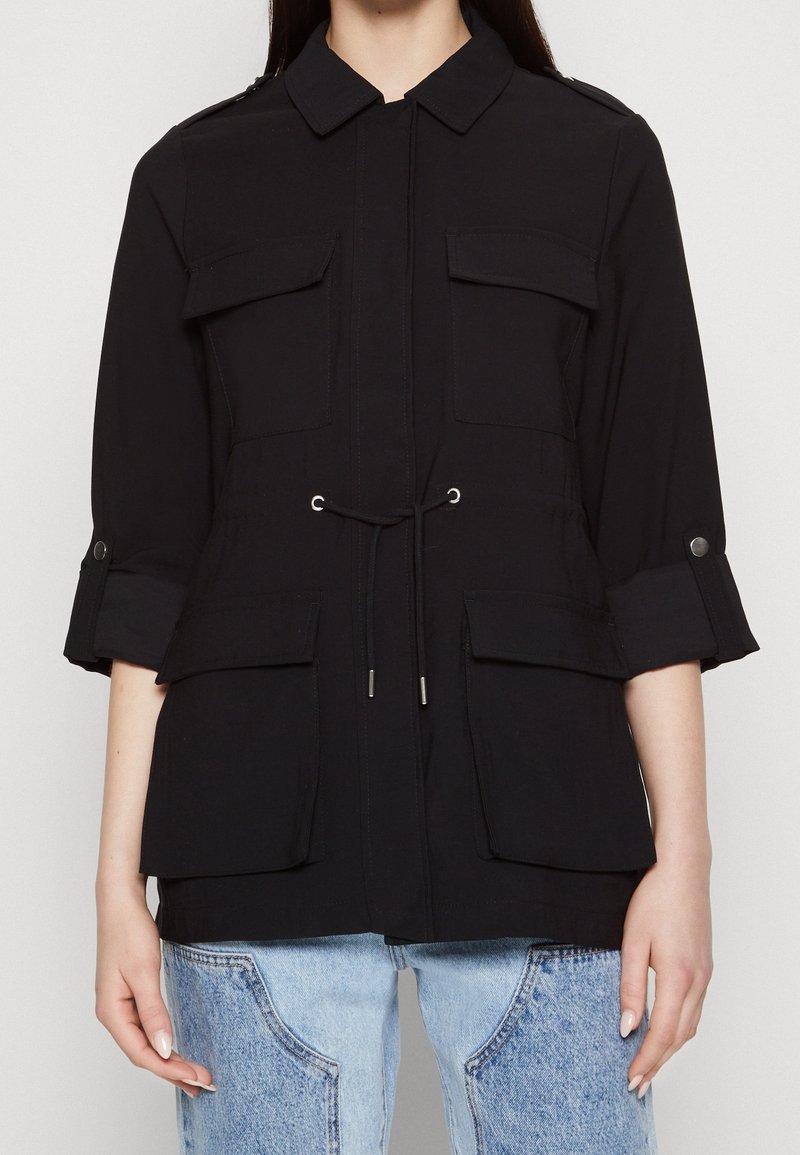 Czarna kurtka robocza z kołnierzykiem, przednimi kieszeniami, regulowanym ściągaczem w talii i podwiniętymi rękawami. Wykonana z lekkiego materiału.