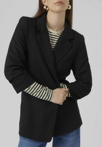Blazer noir ajusté avec des épaules structurées, porté sur un haut à rayures crème et noir. Les mains du mannequin sont jointes à la taille, mettant en valeur des bijoux en or.