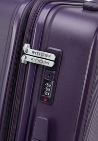 Valigia rigida viola con cerniere argentate, dotata di lucchetto a combinazione e etichette brandizzate. Superficie testurizzata e dettagli di design eleganti.